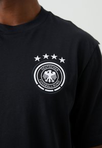 Μαύρο βαμβακερό μπλουζάκι με κεντητό λογότυπο σε κυκλική μορφή, με διπλό δακτύλιο, έναν αετό και πέντε αστέρια από πάνω.