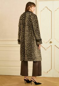 Fekete és bézs foltos hosszú kabát laza szabással, fekete gallérral és ujjmandzsettával, barna nadrágra és fekete magassarkú cipőre viselve.