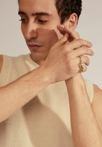 Beige gebreide mouwloze top, close-up van handen die gouden ringen dragen, met nadruk op ingewikkelde details en textuur. Zacht licht benadrukt de kenmerken.