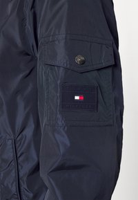 Mørkeblå dunjakke med en blank finish, der har en patch-lomme på ærmet, mærket med et rektangulært "TOMMY HILFIGER" logo.