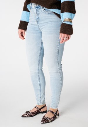 Persoon die lichtblauwe skinny jeans draagt, een bruin-blauw gestreepte trui, puntige ballerina's met luipaardprint en gespen, en zilveren armbanden en ringen.