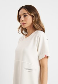 Krémefarbenes Kurzarmkleid mit Rundhalsausschnitt, das eine strukturierte Oberfläche aufweist und auf der Vorderseite ein graues Logo aufgedruckt hat, mit einem roten Etikett am Ärmel.