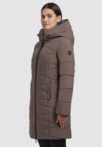 Gevoerde winterjas in bruin met een hoge kraag en een capuchon. Kenmerkt zich door een diagonaal doorgestikt patroon, een rits aan de voorkant en zijzakken.