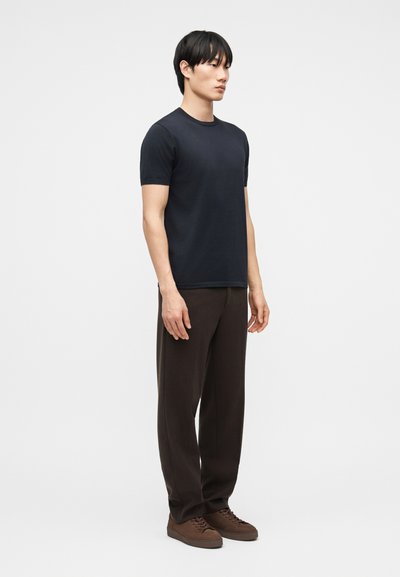 Modèle masculin debout de profil portant un t-shirt noir à manches courtes, un pantalon marron foncé et des baskets marron sur un fond uni.