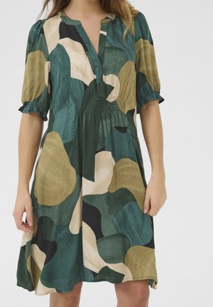 Kobieta ubrana w sukienkę do kolan z krótkimi bufkami i abstrakcyjnym wzorem w zielone, beżowe, czarne i brązowe odcienie.