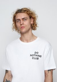 White cotton t-shirt featuring a black embroidered "DO NOTHING CLUB" logo on the left chest. Crew neckline, short sleeves, relaxed fit.
