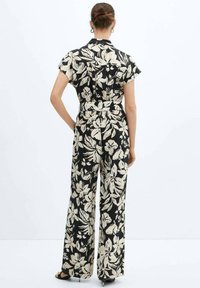 Jumpsuit negro con estampado floral que cuenta con mangas cortas, una cintura ceñida con cinturón y pantalones de pernera ancha con un patrón de flores color crema. Textura de tela suave.