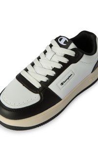 White and black low-top sneaker with white laces, Champion logo on the side and tongue, and beige sole with black tread.