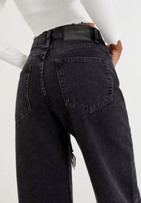 Jean en denim noir taille haute avec une finition délavée, poches arrière visibles et une étiquette texturée. Un léger effilochage est observé à l'ourlet.