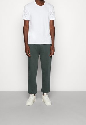 Man draagt een eenvoudige witte korte mouwen t-shirt, donkergroene joggingsbroek en witte sneakers tegen een neutrale achtergrond.