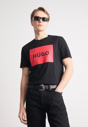 Black cotton t-shirt featuring a bold red rectangular graphic with "HUGO" printed in black. Worn with dark jeans and black sunglasses.