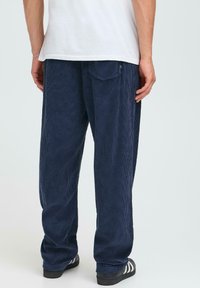 Pantalons en velours côtelé bleu marine à rayures verticales, coupe décontractée, poches latérales et arrière, assortis à des chaussures à rayures noires et blanches.