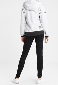 Lys grå vandtæt jakke med hætte, sorte accenter og "SUPERDRY" logo. Sammen med tætsiddende sorte jeans og hvide sneakers.