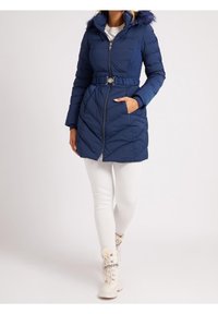 Mujer con un abrigo acolchado azul marino con cinturón y capucha con borde de piel, mallas blancas y botas de invierno blancas con cordones.
