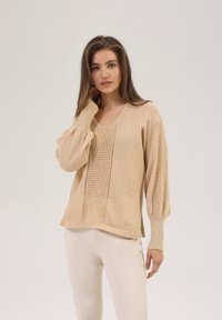 Pull beige tricoté avec un décolleté en V, présentant des sections texturées et des manches ballons. Tissu doux et léger avec des poignets côtelés.