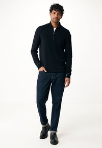 Felpa nera con zip e maniche lunghe, abbinata a jeans tapered blu scuro e stivaletti neri alla caviglia. Presenta polsini e orlo a costine.