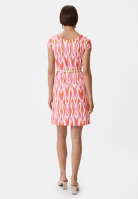 Das kurzärmlige Kleid hat ein lebhaftes, pinkes, oranges und weißes abstraktes Muster, wird in der Taille mit einem Gürtel gerafft und hat eine Länge bis oberhalb des Knies.