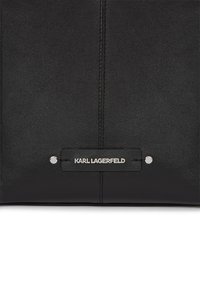 Sac en cuir noir avec une texture lisse, un design minimaliste et une plaque logo en métal argenté avec deux clous en bas.