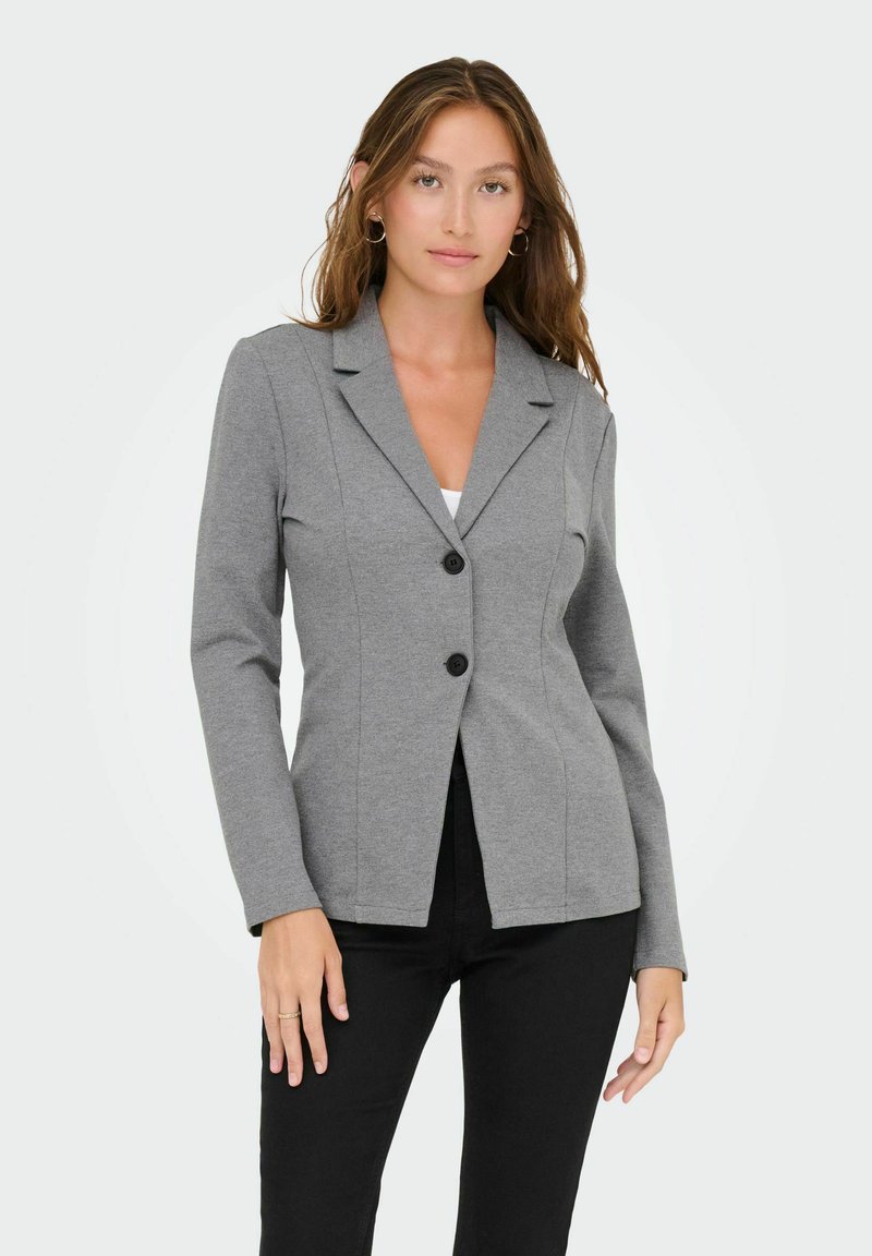 Femme portant un blazer gris ajusté avec des boutons noirs sur un haut blanc, associée à un pantalon noir, debout devant un fond clair uni.