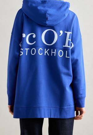 Osoba ubrana w jasnoniebieską bluzę z kapturem z białym napisem "STOCKHOLM" i częściowo dużymi białymi literami na plecach, w połączeniu z ciemnymi spodniami.