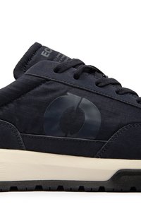 Gros plan d'une basket noire avec des lacets noirs, un logo circulaire foncé avec des flèches sur le côté et une semelle intermédiaire blanche avec une semelle extérieure noire.