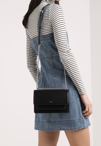 Žena v proužkovaném topu s dlouhým rukávem a džínových šatech nese černou koženou kabelku s stříbrným řetízkovým popruhem přes rameno.