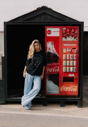 Automat sprzedający z czerwonym zewnętrzem z brandingiem Coca-Coli, otoczony różnymi wyborami napojów. Model stoi w luźnych jasnoniebieskich spodniach i czarnej koszulce.