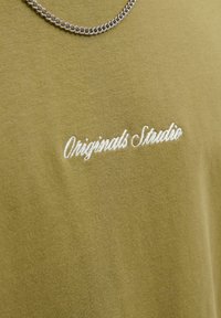 Alyvuogių spalvos audinys su balta siuvinėta užrašu "Originals Studio" ir sidabrinė grandinėlė, gulinti virš užrašo.