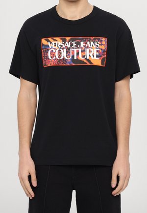 T-shirt noir à manches courtes avec un patch rectangulaire imprimé animalier orange et bleu affichant "Versace Jeans Couture" en texte blanc sur la poitrine.