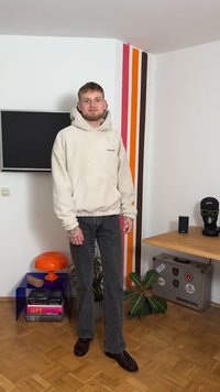 Béžová fleecová mikina s kapucí a přední kapsou, šedé džínsy s širokými nohavicemi a hnědé boty. Interiér zdobí barevný pruhovaný válec a rostliny.