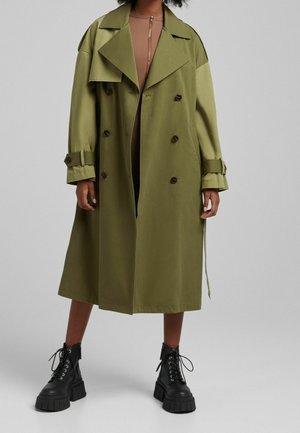 Persona che indossa un trench coat over-size verde oliva, una giacca marrone con zip e stivaletti neri chunky con lacci, in piedi contro uno sfondo chiaro e neutro.