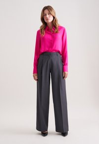 Fuchsia zijden shirt met knopen aan de voorkant, gecombineerd met hoge grijze broek met streepjes. Zwarte hakken maken de look af.