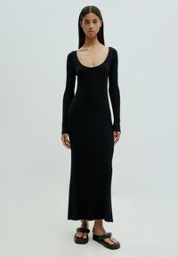 Schwarzes geripptes Strickkleid mit Rundhalsausschnitt und langen Ärmeln, das bis zu den Knöcheln reicht. Kombiniert mit schwarzen flachen Sandalen. Glatte Textur.