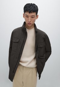 Giacca button-up marrone scuro con colletto alto e due tasche frontali, indossata sopra un pullover a coste beige chiaro e pantaloni beige.