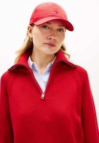 Κόκκινο πουλόβερ με φερμουάρ, με ριμπ λαιμόκοψη και πλεκτή υφή, φορεμένο πάνω από ένα ανοιχτό μπλε ριγέ πουκάμισο. Συνδυάζεται με κόκκινο καπέλο μπέιζμπολ.