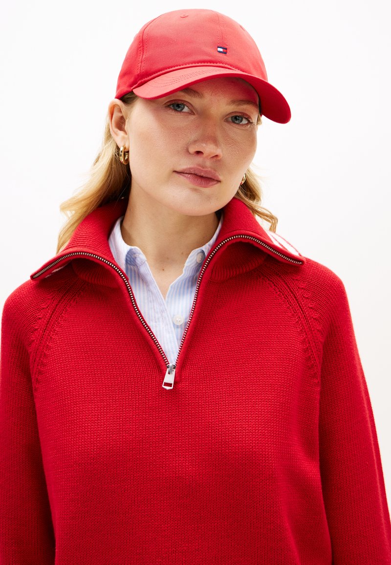 Κόκκινο πουλόβερ με φερμουάρ, με ριμπ λαιμόκοψη και πλεκτή υφή, φορεμένο πάνω από ένα ανοιχτό μπλε ριγέ πουκάμισο. Συνδυάζεται με κόκκινο καπέλο μπέιζμπολ.