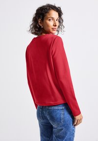 Czerwona bluzka z długim rękawem i okrągłym dekoltem. Gładka faktura, luźny krój, w połączeniu z jasnoniebieskimi dżinsami z tylnymi kieszeniami.