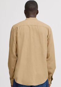 Langermet skjorte i lys tan, laget av myk stoff. Har bakre yoke, knappeender og en avrundet sømkant. Enkel og minimalistisk design.