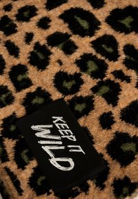 Tessuto con stampa leopardata nelle tonalità di marrone presenta macchie nere e verdi. Un'etichetta nera con la scritta "KEEP IT WILD" in cucitura bianca è ben visibile.