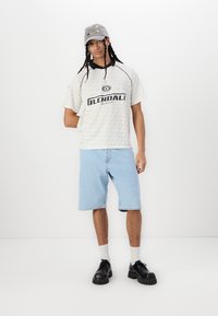 Short-sleeve, light-colored shirt with black text, paired with mid-length light blue denim shorts. Black chunky shoes and a grey cap.