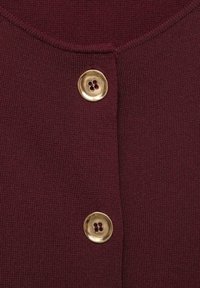 Měkká pletenina v barvě kaštanové, detail včetně kulatého zlatého knoflíku se čtyřmi otvory, který zdůrazňuje strukturu látky a detaily knoflíku.