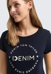 Tamsiai mėlynas medvilninis marškinėlis su apvaliu baltu tekstu, kuriame parašyta "DENIM TOM TAILOR" su širdelės akcentu, apvaliu kaklu ir trumpomis rankovėmis.