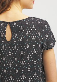 Mujer con una blusa de manga corta negra con un patrón geométrico en blanco y verde azulado, y un pequeño cierre en forma de ojales en la parte superior de la espalda.
