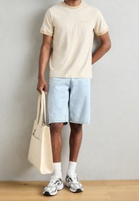 Beige bomulds t-shirt med kontrastfarvet krave, lyseblå denimshorts, hvide sokker og grå træningssko, der holder en beige mulepose.