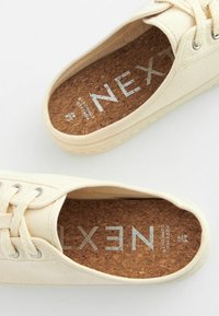 Baskets en toile couleur écru, avec une semelle en liège ornée de texte argenté, des lacets ronds et une semelle en caoutchouc texturée.