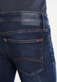 Mörkblå denimjeans med en läderetikett märkt "TOMMY JEANS." Har kontrasterande sömmar och en bakfickdesign med broderad detalj.