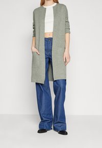 Lång, öppen cardigan i ljusgrön stickad tyg med trekvartslånga ärmar, i kombination med högmidjade blå vida jeans och svarta skor.