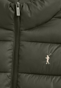 Närbild av en grön vadderad jacka med central dragkedja och broderad beige polo-spelarlogotyp på höger sida.