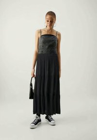 Svart maxi-kjole med en tettsittende denim-overdel, spaghetti-stropper og flytende, lagdelt skjørt. Brukt med svarte sneakers og bærende en svart håndveske.
