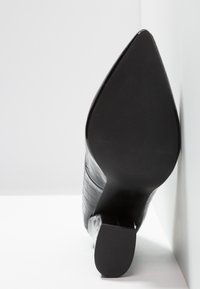 Chaussure à talons hauts en cuir verni noir avec un bout pointu, présentant un motif crocodile texturé et une semelle lisse.